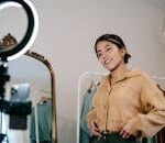 Low angle of positive ethnic female blogger shooting video on cellphone on tripod with shining lamp